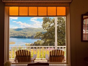 Une pièce dotée d’une grande fenêtre donnant sur un lac et des collines boisées. On y trouve deux chaises en bois sur un balcon.