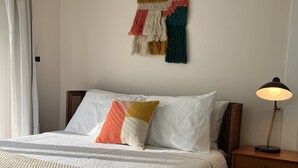 Un lit bien fait, avec une couette blanche et des oreillers, une table de chevet en bois avec une lampe, et une décoration murale au-dessus du lit.