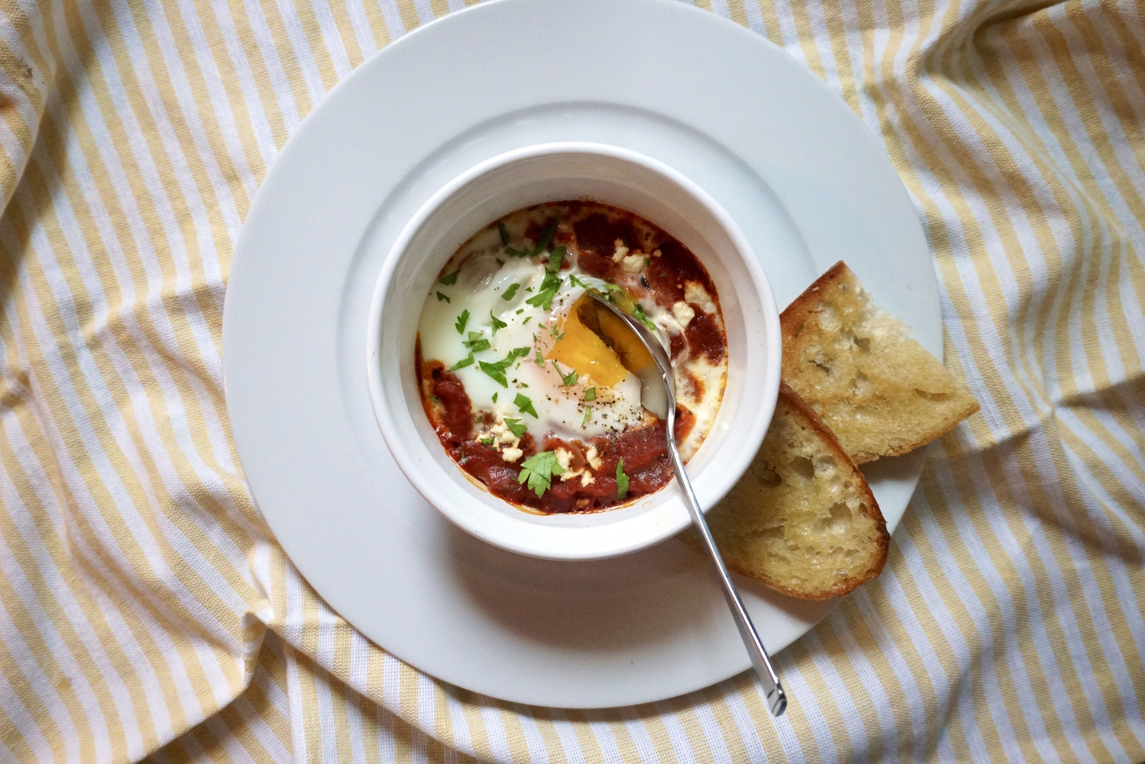 A bowl of soup with a spoon and two pieces of bread on a white plate, set on a striped cloth.