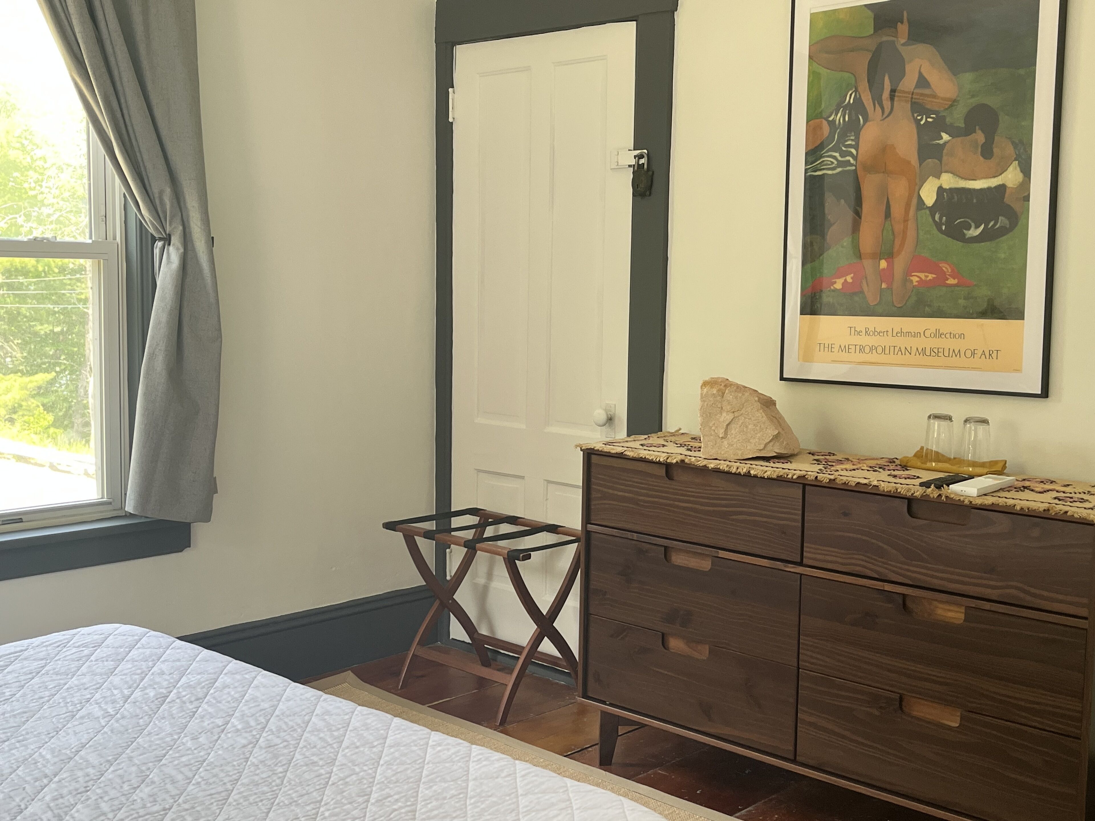 A bedroom with a bed, a dresser, a small stool, a painting, and a window with curtains.