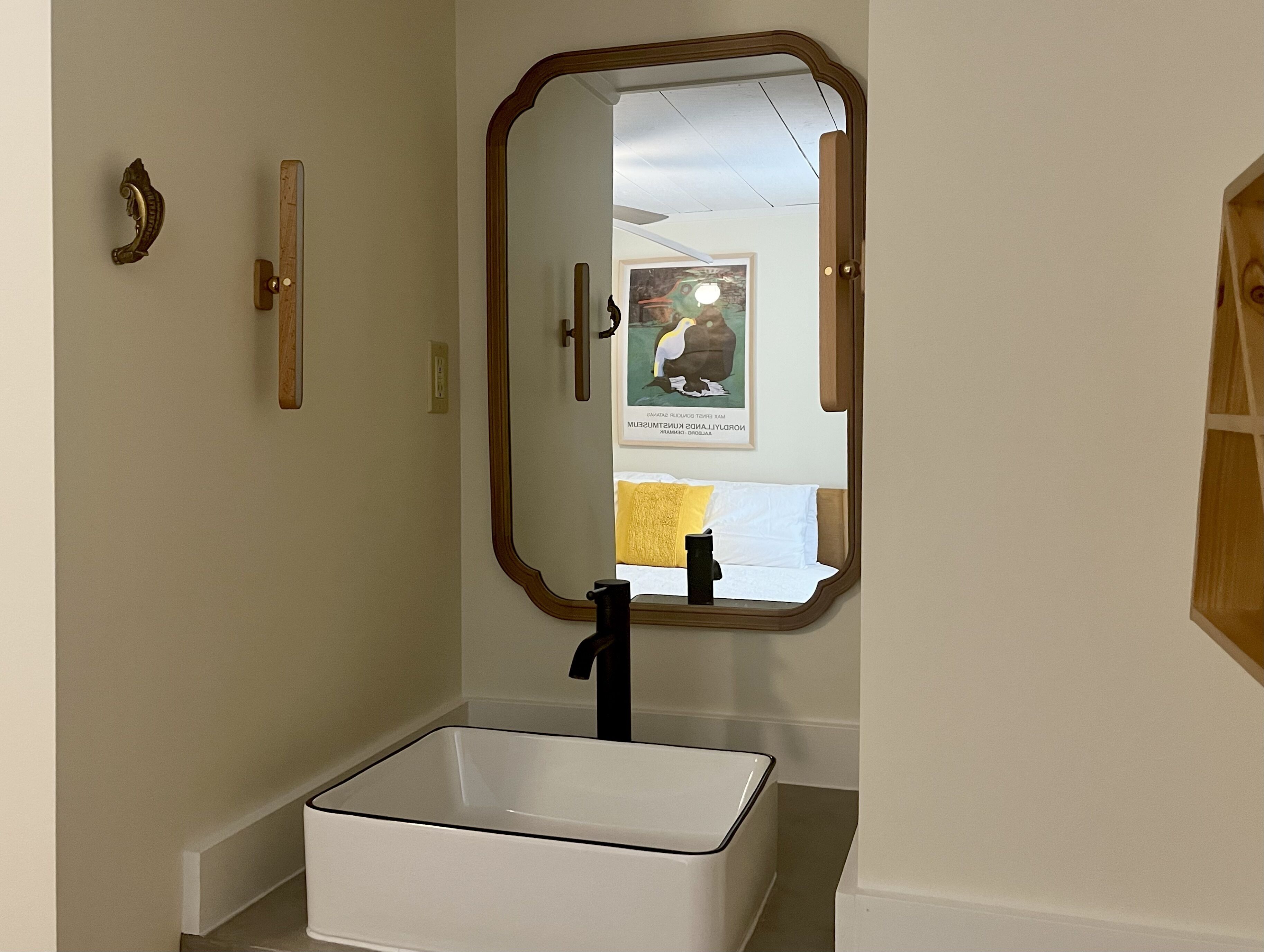 Un baño con lavamanos blanco, un espejo y un pomo de madera.