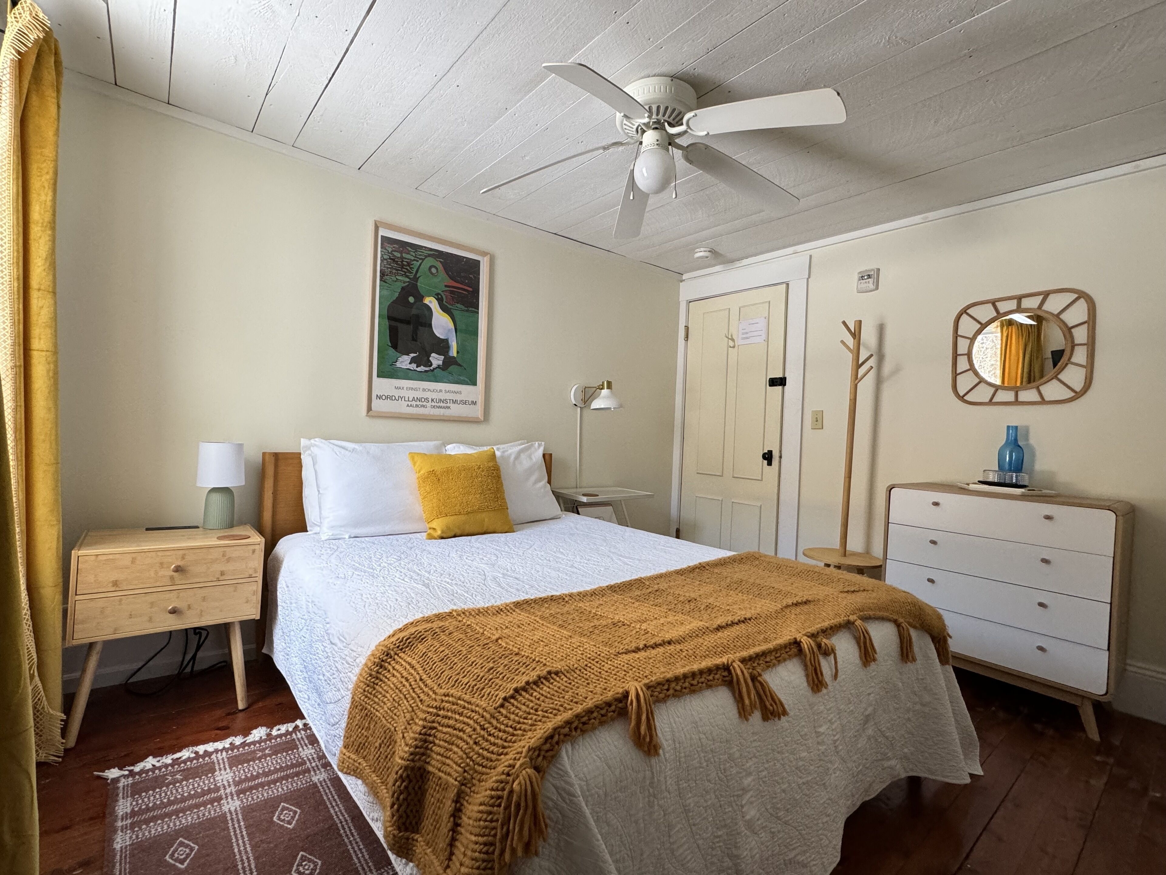A bedroom with a bed, a nightstand, a dresser, a mirror, a ceiling fan, and a painting on the wall.