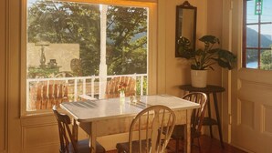 Un coin repas avec une table et des chaises, une fenêtre donnant sur des arbres, une plante en pot et une porte offrant une vue sur une montagne.