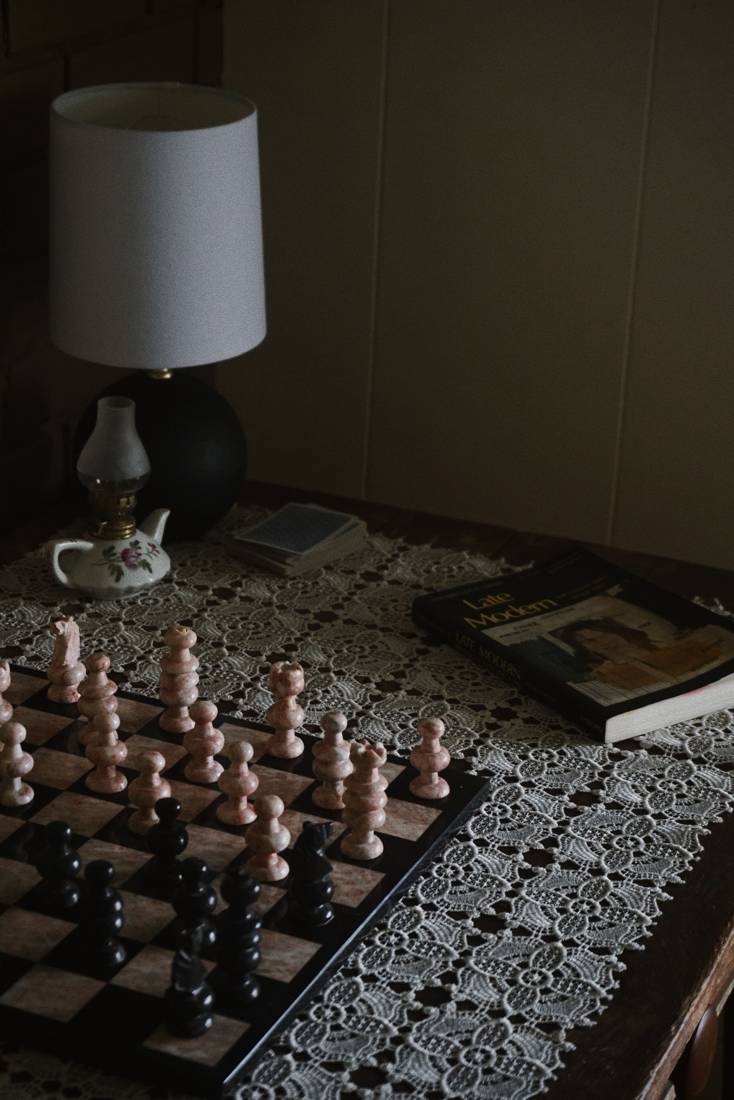 A chessboard on a lace tablecloth with a book and a lamp beside it.