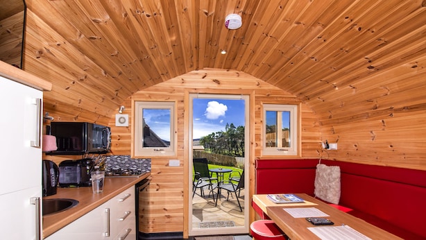 A compact wooden cabin with a kitchenette, a dining area with a red couch, and a view of the outdoors through a door. - Tigh an Each B&B & Laggan Glamping (Newtonmore)