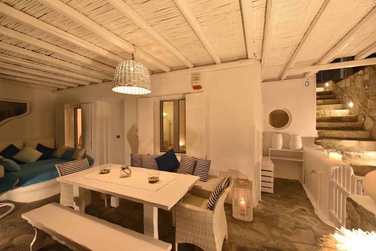 Un interior acogedor con una mesa de comedor blanca, sillas de mimbre, un sofá azul y una escalera de piedra.