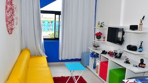 Una sala de estar con un sofá amarillo, una mesa azul, un tapete rojo y un televisor sobre un estante blanco.