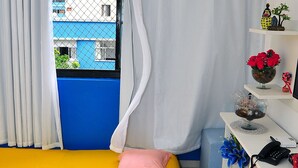 Una habitación con una cama amarilla, cortinas blancas, un tapete rojo y una ventana con vistas a edificios.