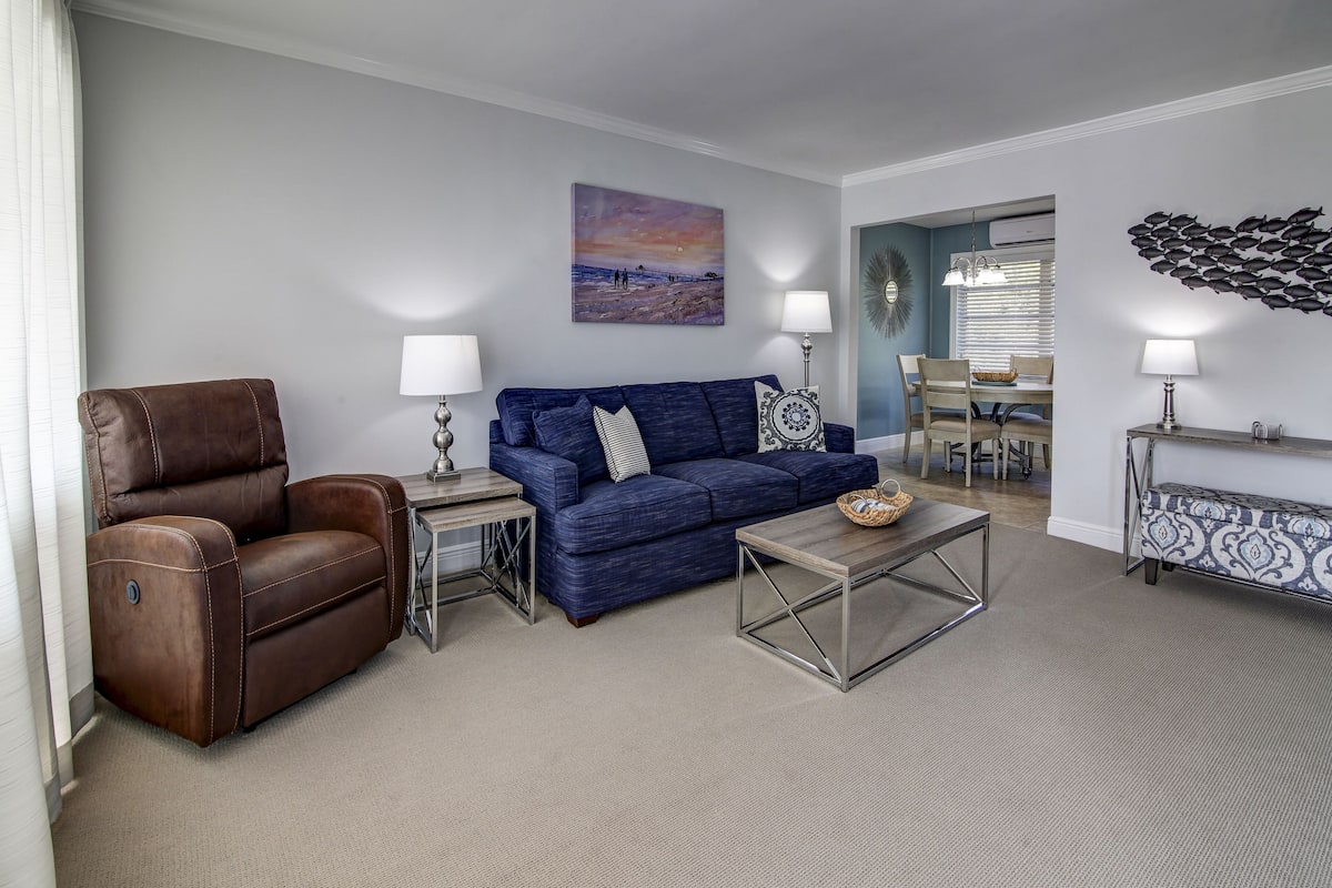 A living room with a brown leather recliner, a blue sofa, a coffee table, and a dining area in the background.