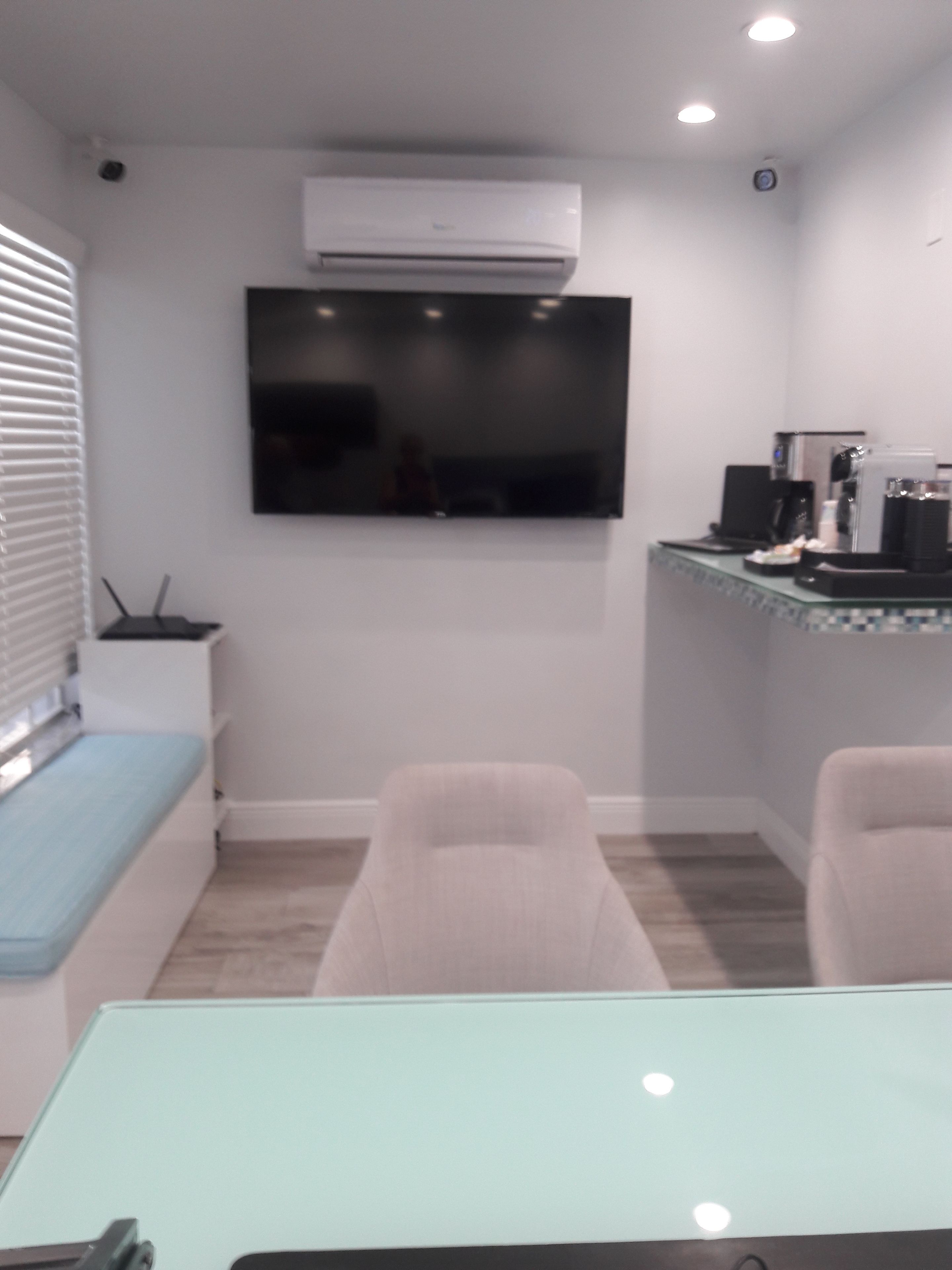 A compact room with a flat-screen TV mounted on the wall, a small seating area with a bench, and a kitchenette with a coffee machine.