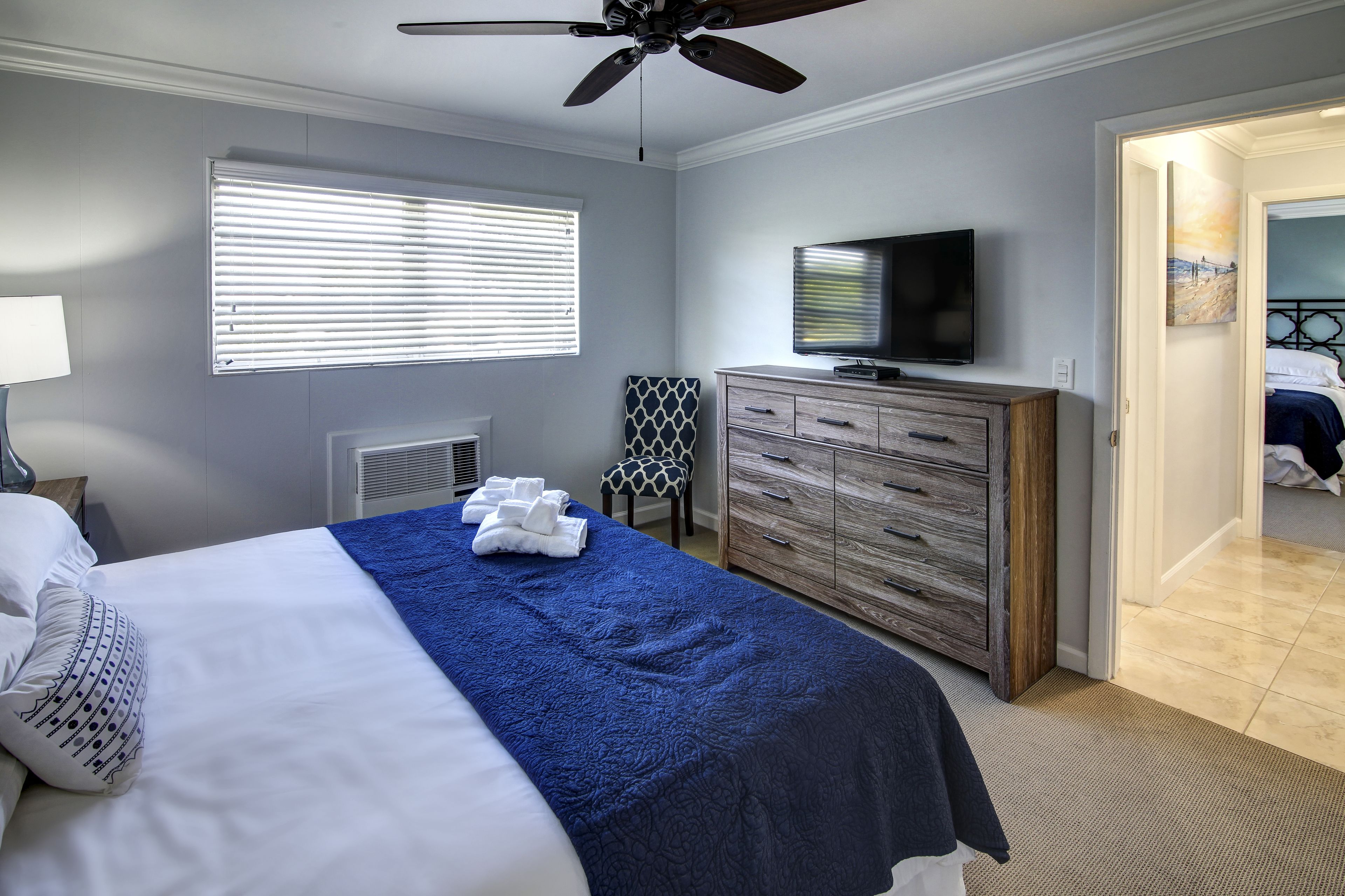Un dormitorio con cama, ventilador de techo, televisor, cómoda y ventana con persianas.