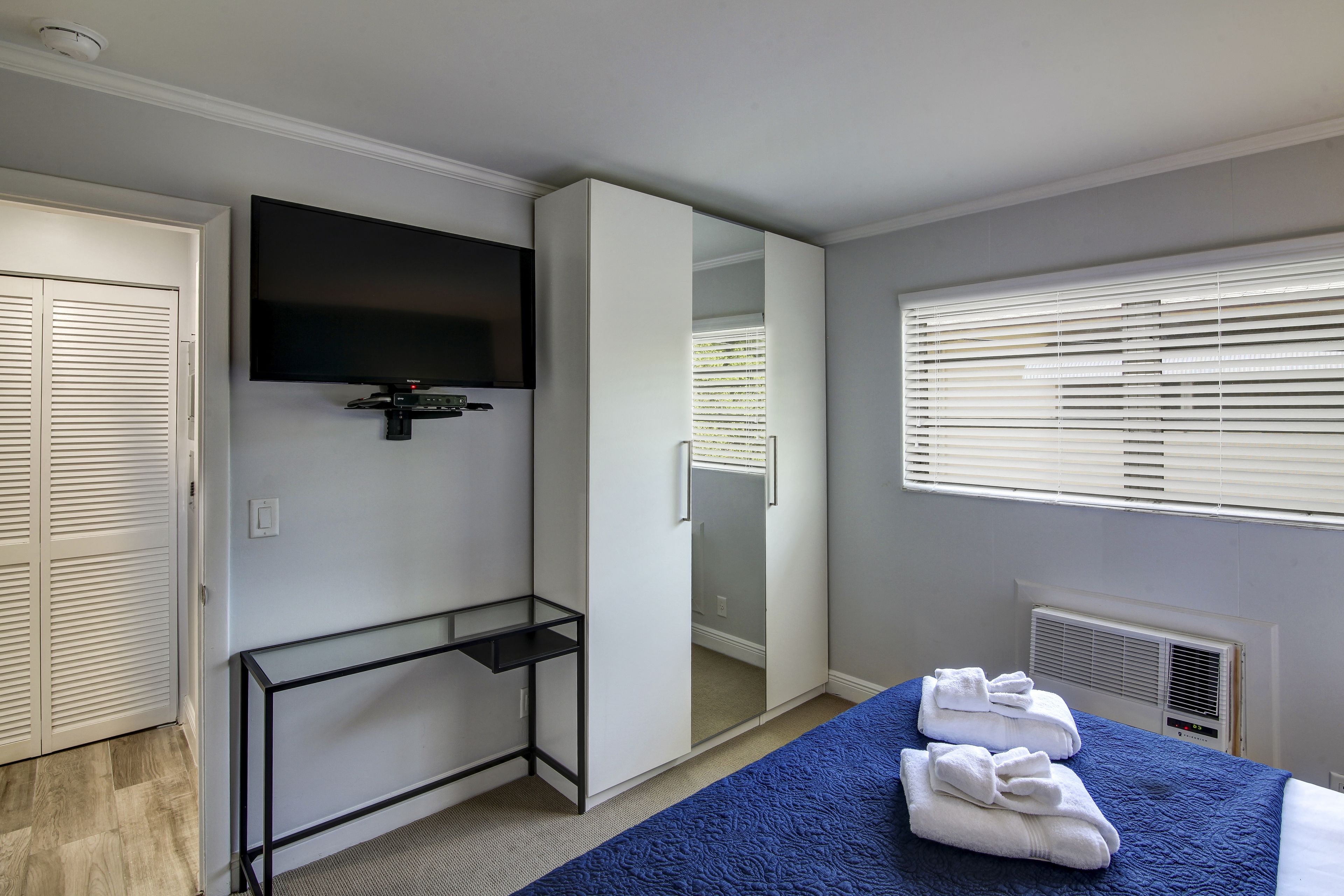 A hotel room with a bed, a TV mounted on the wall, a wardrobe, and a window with blinds.
