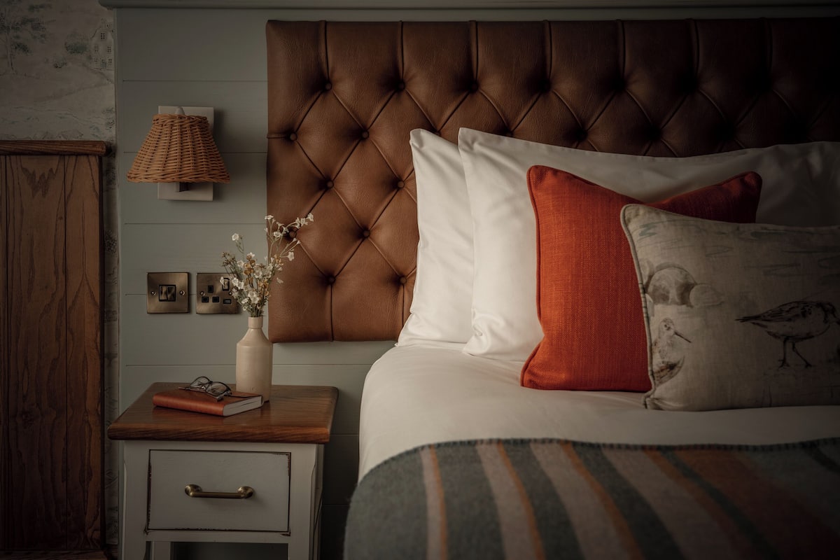Un dormitorio con cabecera capitonada, una mesita de noche con lámpara, un florero con flores y una cama con ropa de cama a rayas.