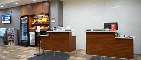 A reception area with wooden counters, a digital screen, a refrigerator stocked with beverages, and a snack display.