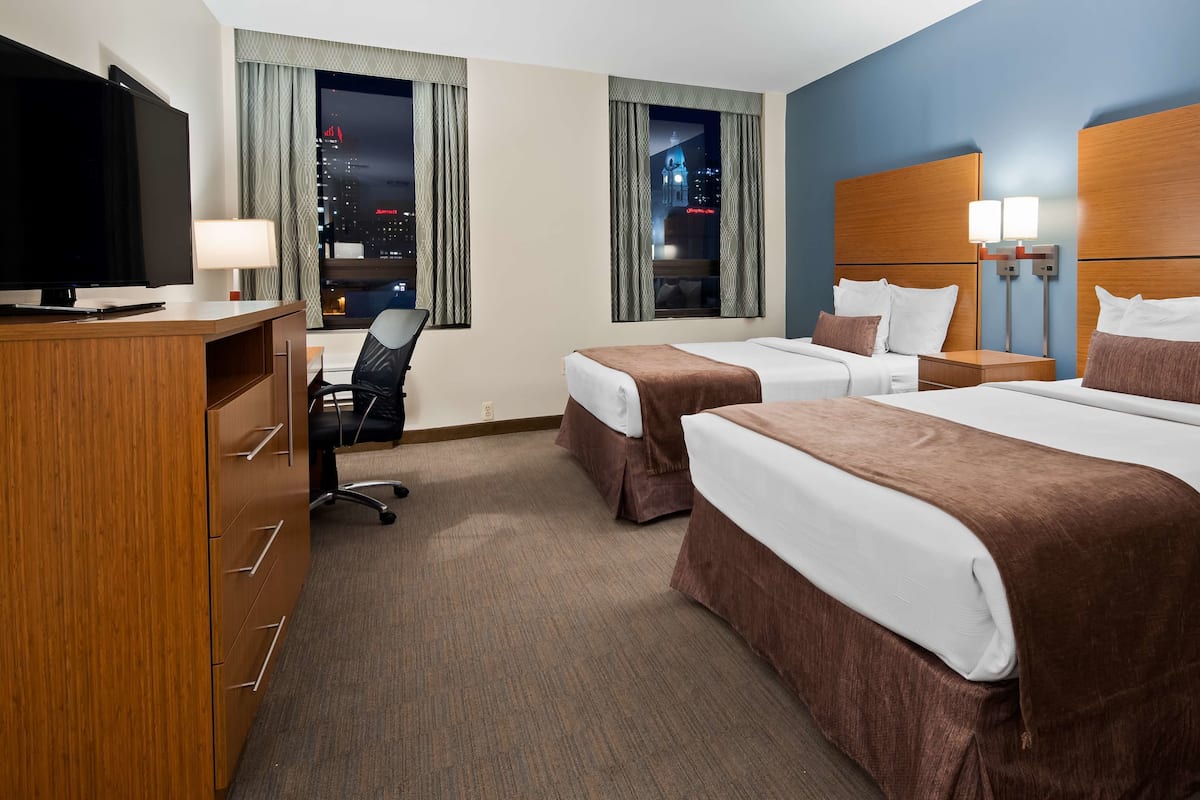 A hotel room with two beds, a desk with a chair, a TV, and a view of a cityscape at night.