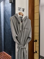 A grey robe with "The Manor" embroidered on it, hanging on a wooden hanger in a hallway with a herringbone-patterned wall and a wooden door.