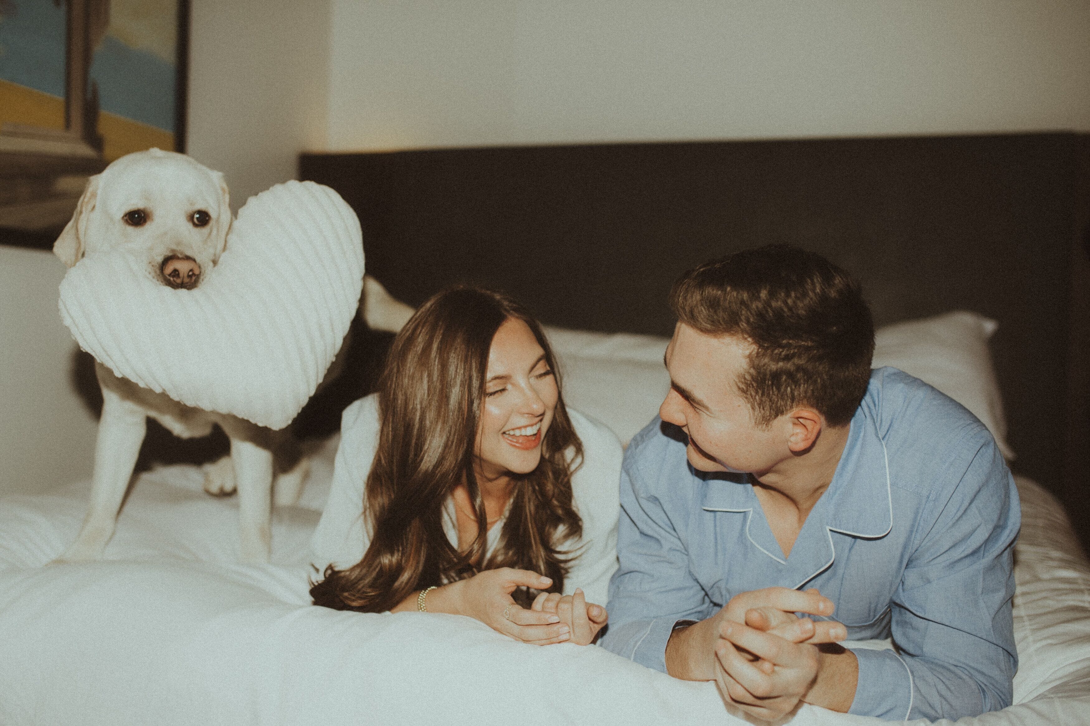 Um casal deitado em uma cama com um cachorro usando uma coleira em formato de coração.
