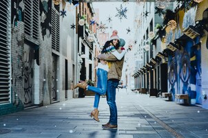 Una pareja se abraza en un estrecho callejón flanqueado por edificios de ladrillo y luces decorativas.