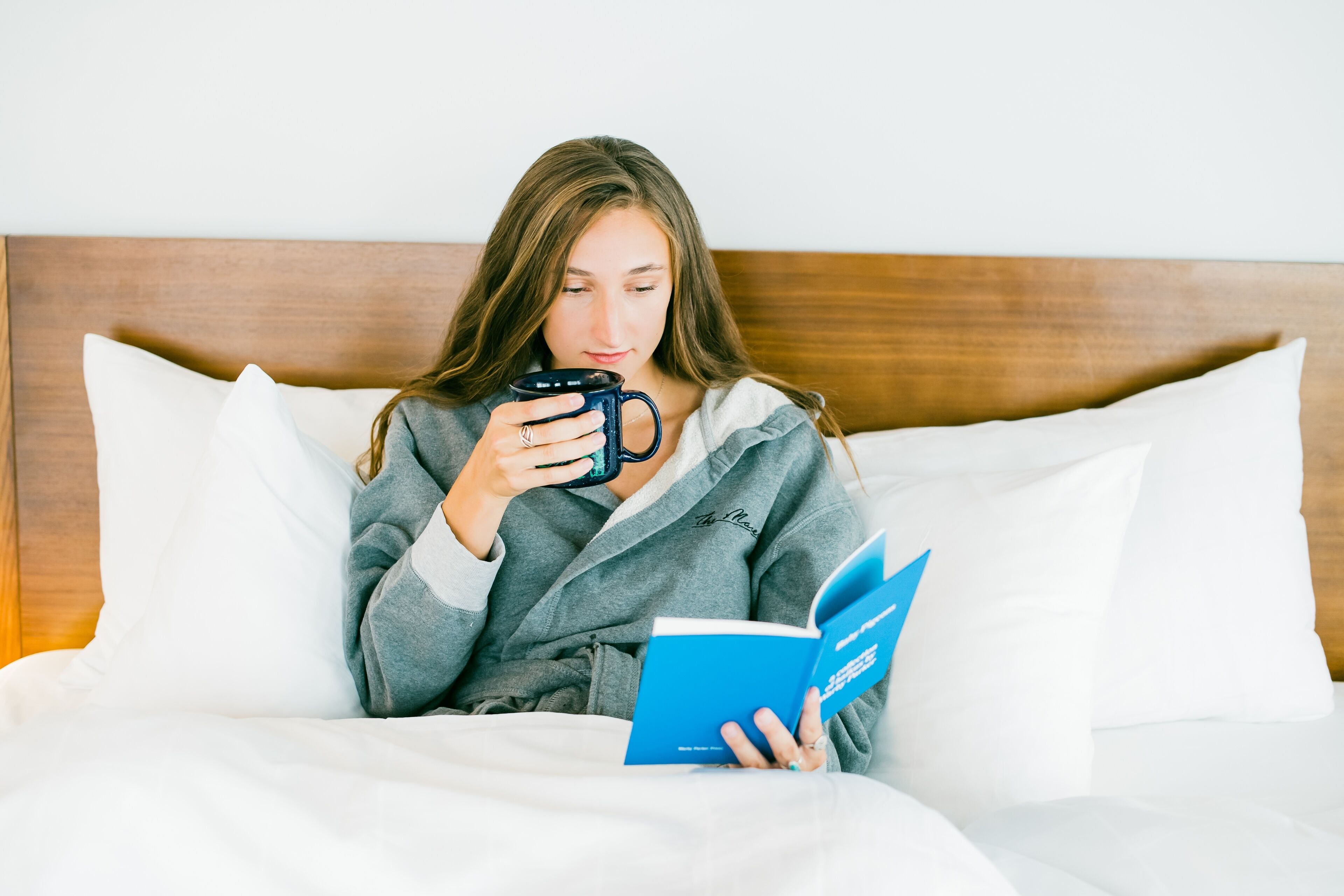 A person sitting on a bed with a book and a mug, in a room with a wooden headboard and white pillows.