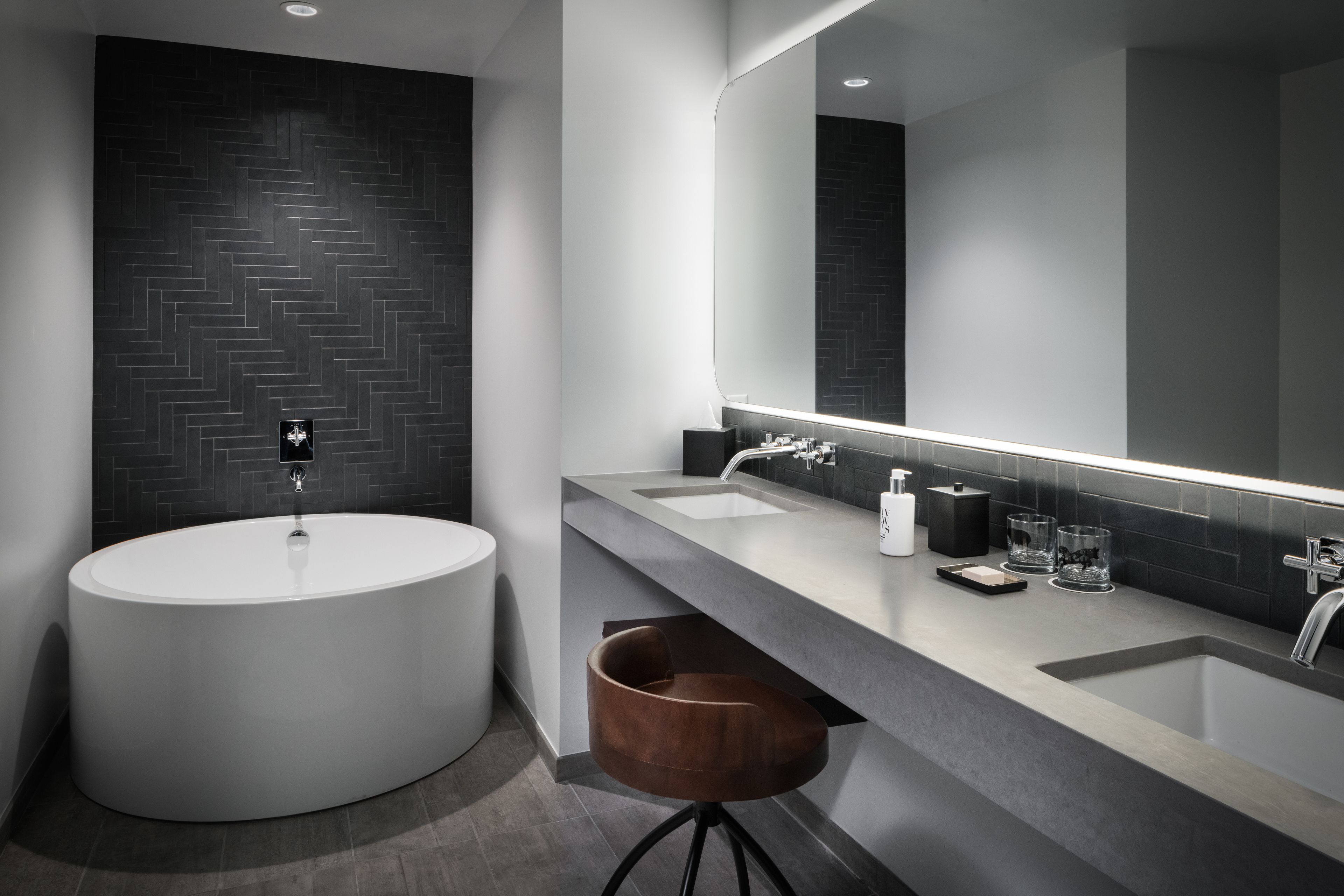 A modern bathroom with a large round bathtub, two sinks, a mirror, and a wooden stool.