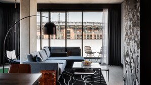 A modern living room with a sofa, a dining table, and a view of a bridge through large windows. - The Maven Hotel at Dairy Block (Denver)