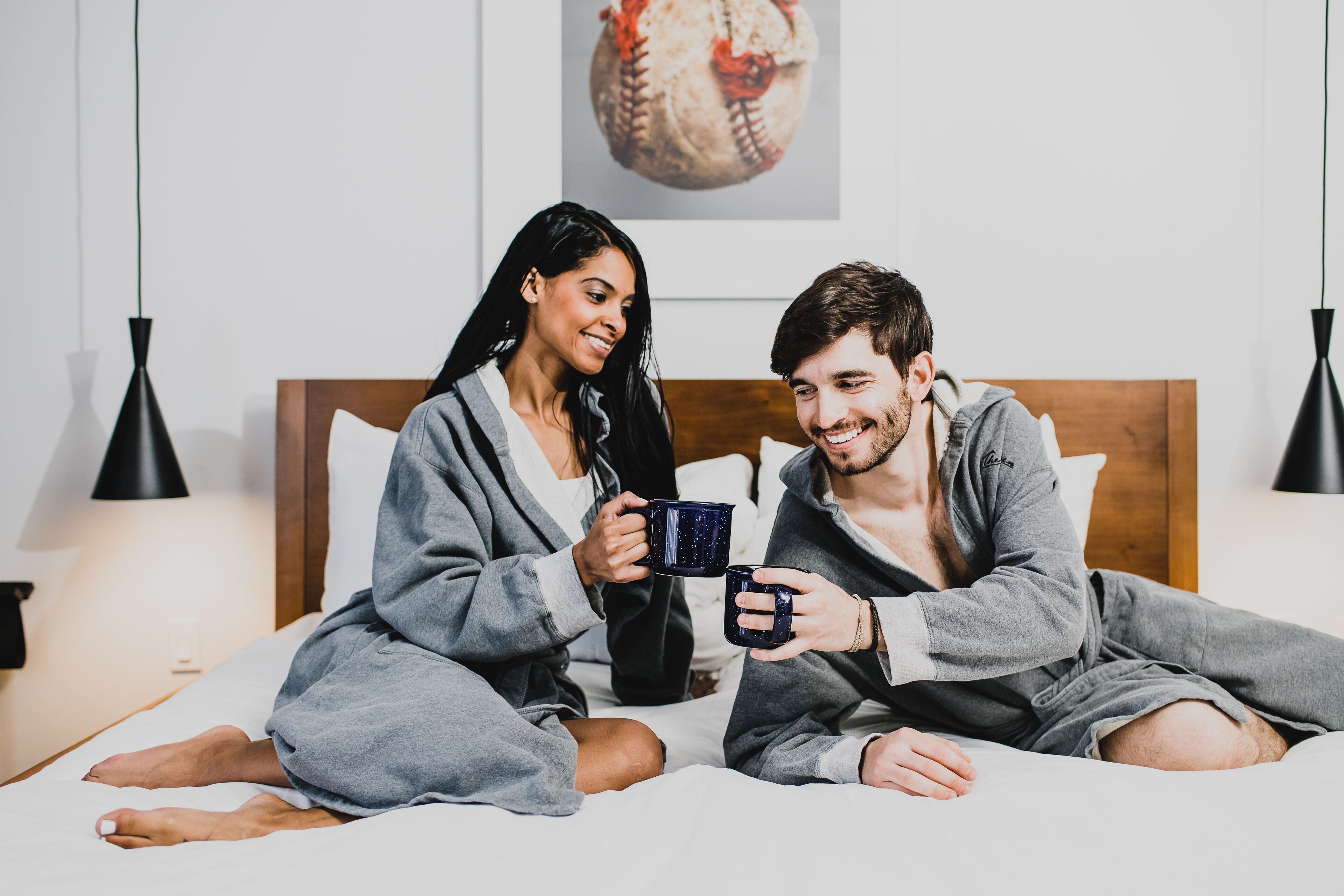 A couple sitting on a bed in a hotel room, holding mugs.