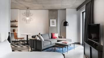 Una sala moderna con sofá, mesa de centro y televisor. Hay un comedor con mesa y sillas. La habitación tiene techo de concreto y un ventanal grande.