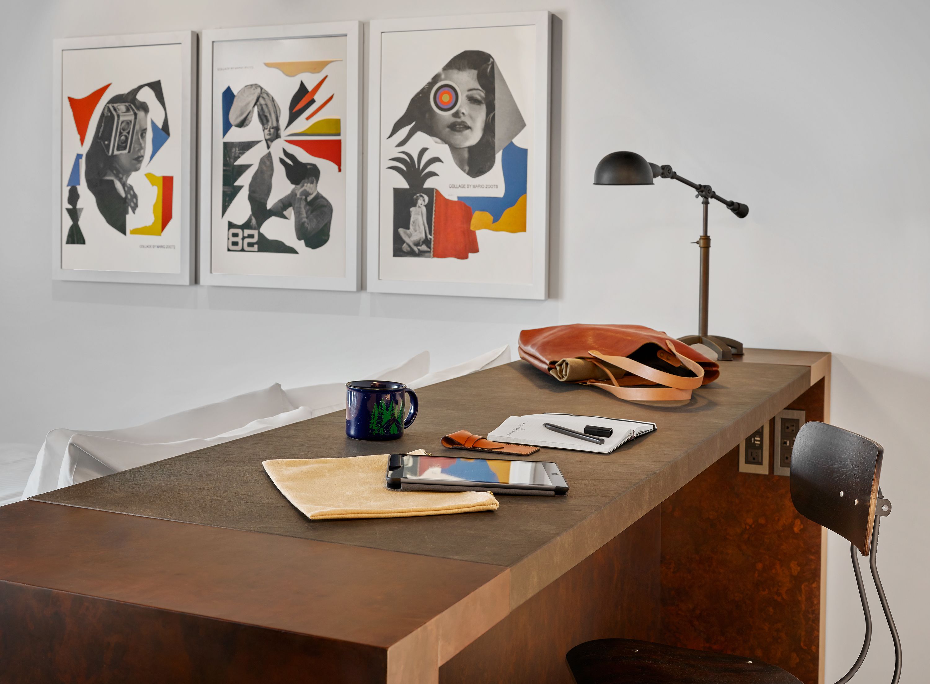 A wooden desk with a laptop, a notebook, a pen, a mug, and a handbag. Three framed artworks are hung on the wall above the desk.