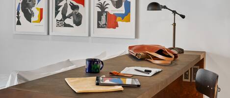 A wooden desk with a laptop, a notebook, a pen, a mug, and a handbag. Three framed artworks are hung on the wall above the desk.