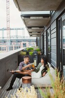 Una pareja sentada en un banco acolchado en una terraza azotea con vistas a la ciudad, suelo de hormigón y barandilla de metal.