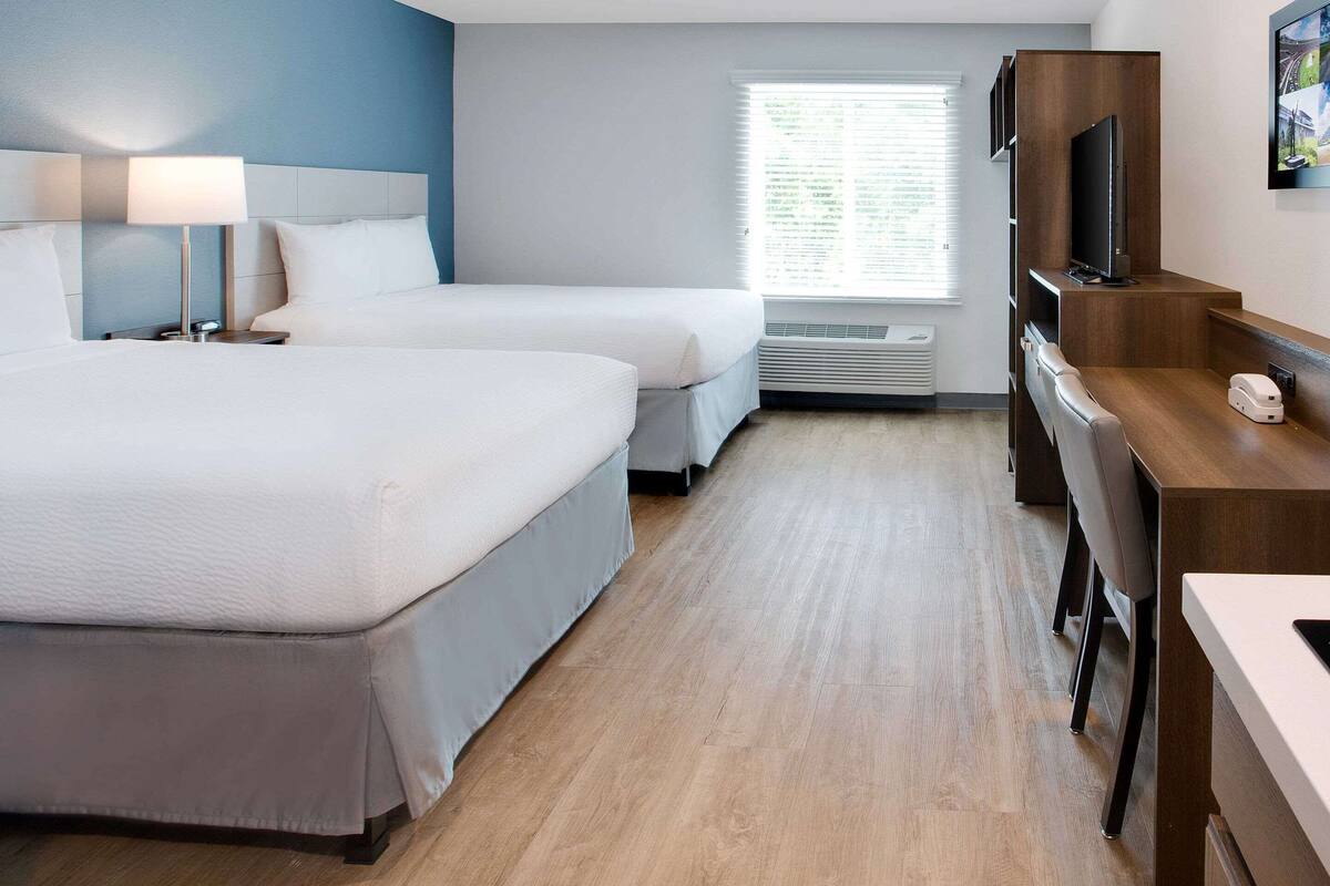 A hotel room with two beds, a desk with a flat-screen TV, a lamp, and a window with blinds.