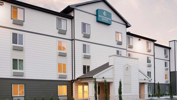 Il s’agit d’un hôtel à plusieurs étages, sur lequel on peut lire l’inscription « Woodspring Suites ». Il présente un design moderne, avec un toit plat, de grandes fenêtres et une façade en pierre à l’entrée.