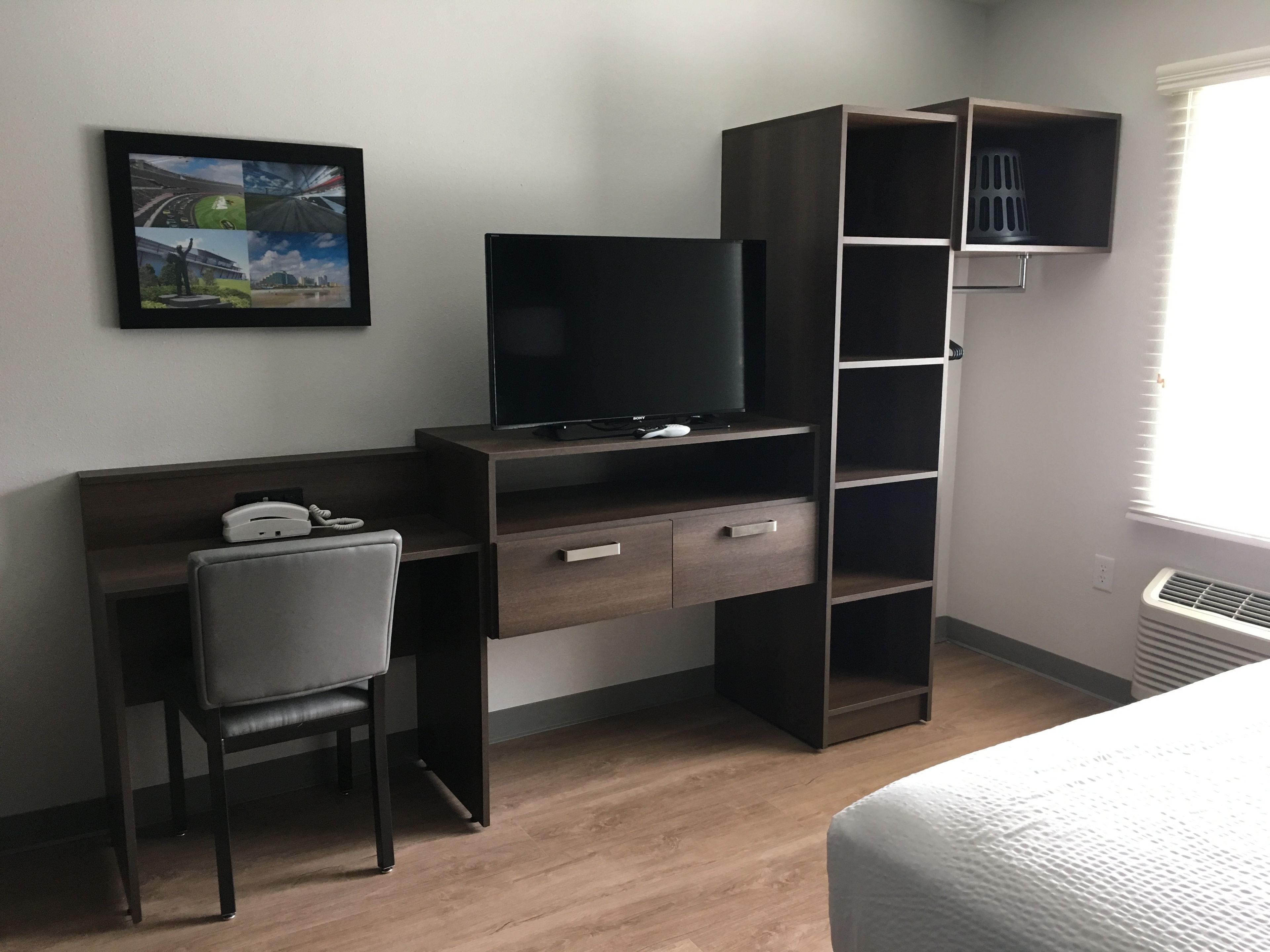 Une chambre d’hôtel avec un bureau en bois, une chaise, un téléviseur à écran plat, une étagère et un tableau encadré au mur.