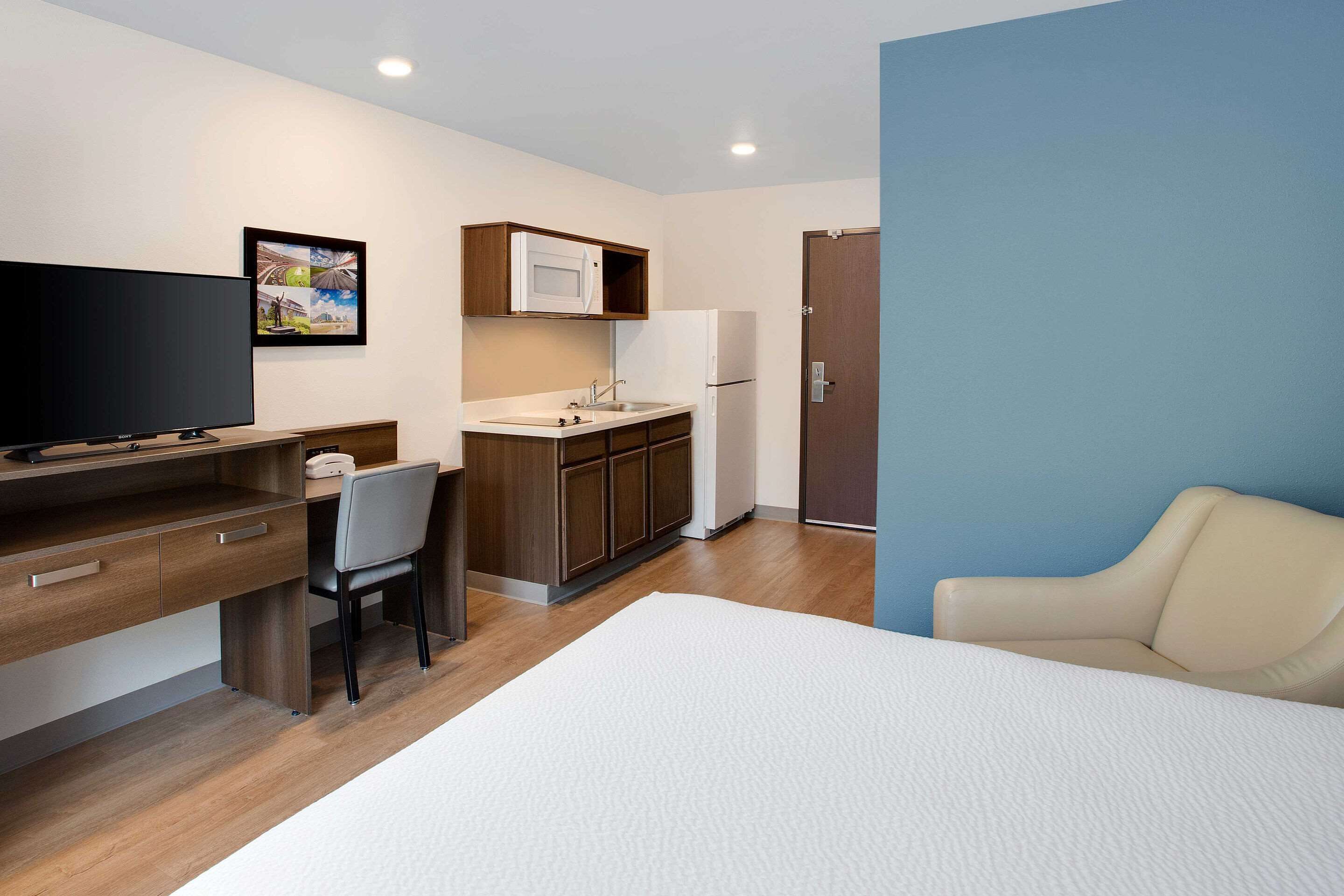 A hotel room with a bed, a desk with a chair, a kitchenette with a microwave, and a door.