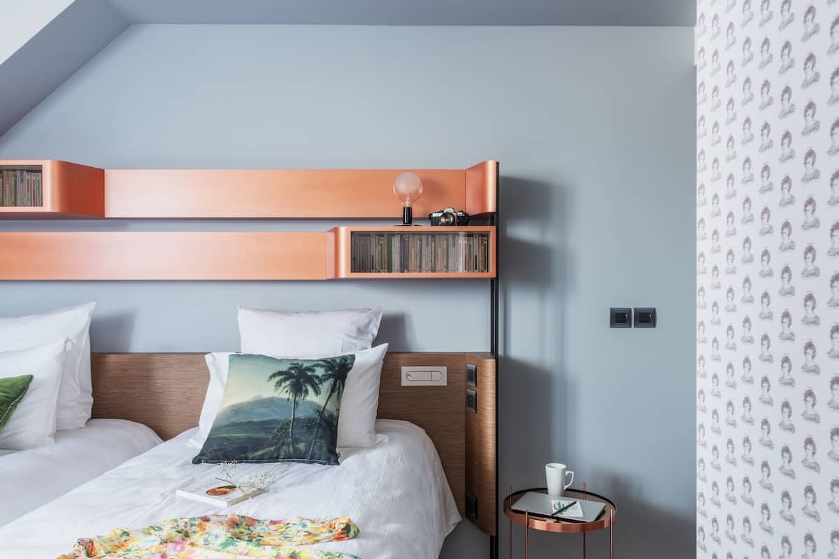 Ein Hotelzimmer mit einem Bett, einem Nachttisch, einer an der Wand befestigten Ablage mit Büchern und einer Kamera sowie einer Wandtapete mit Muster.
