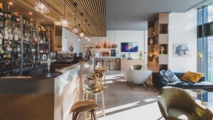 A modern bar area with a wooden ceiling, bar stools, and a variety of bottles. There is a seating area with a sofa and armchairs, and a television mounted on the wall. - Golden Tulip Saint Malo - Le Grand Bé (Saint-Malo)