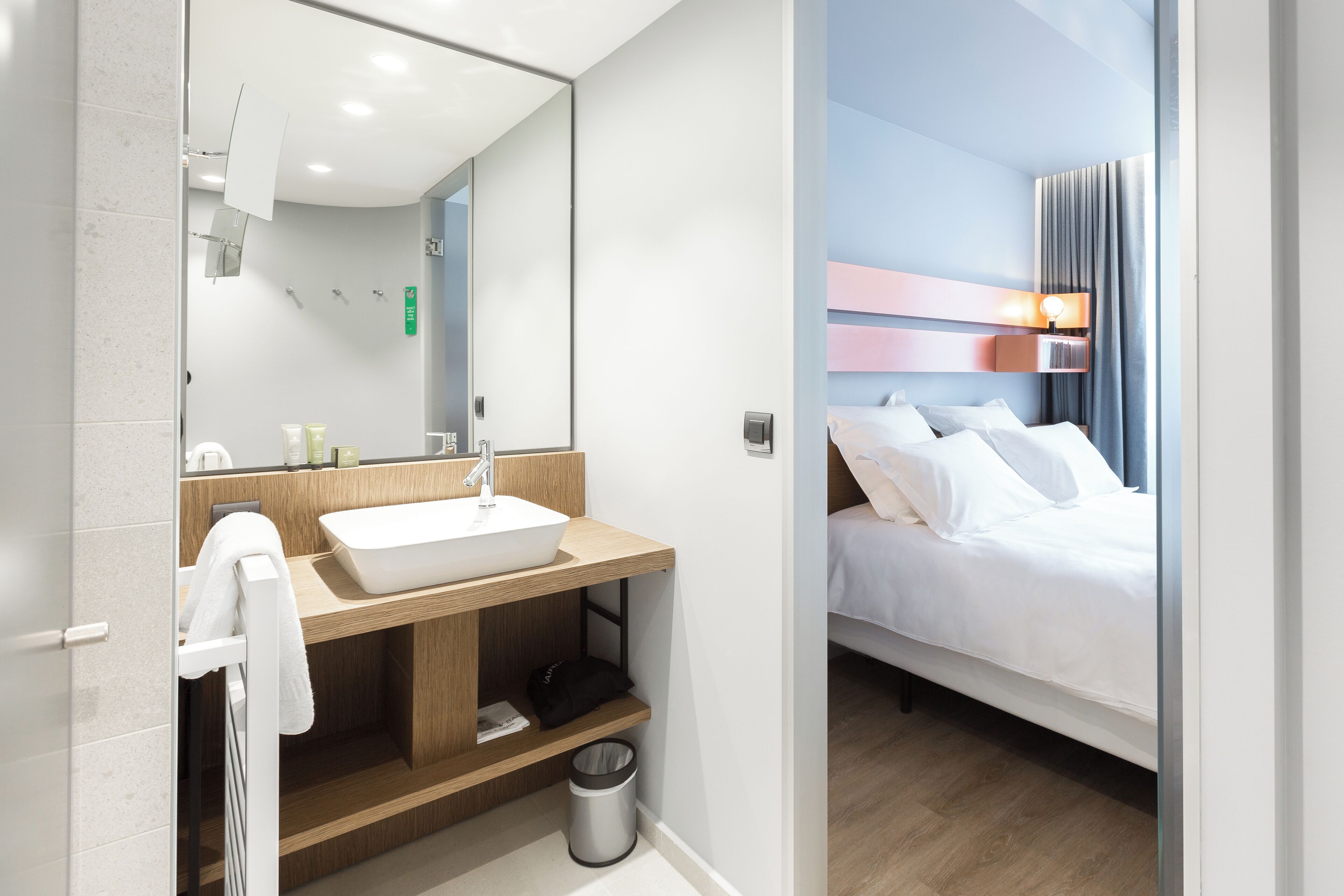 A modern hotel room with a large mirror, a wooden vanity with a white sink, a bed with white linens, and a nightstand with a lamp.