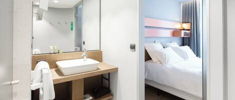 A modern hotel room with a large mirror, a wooden vanity with a white sink, a bed with white linens, and a nightstand with a lamp.