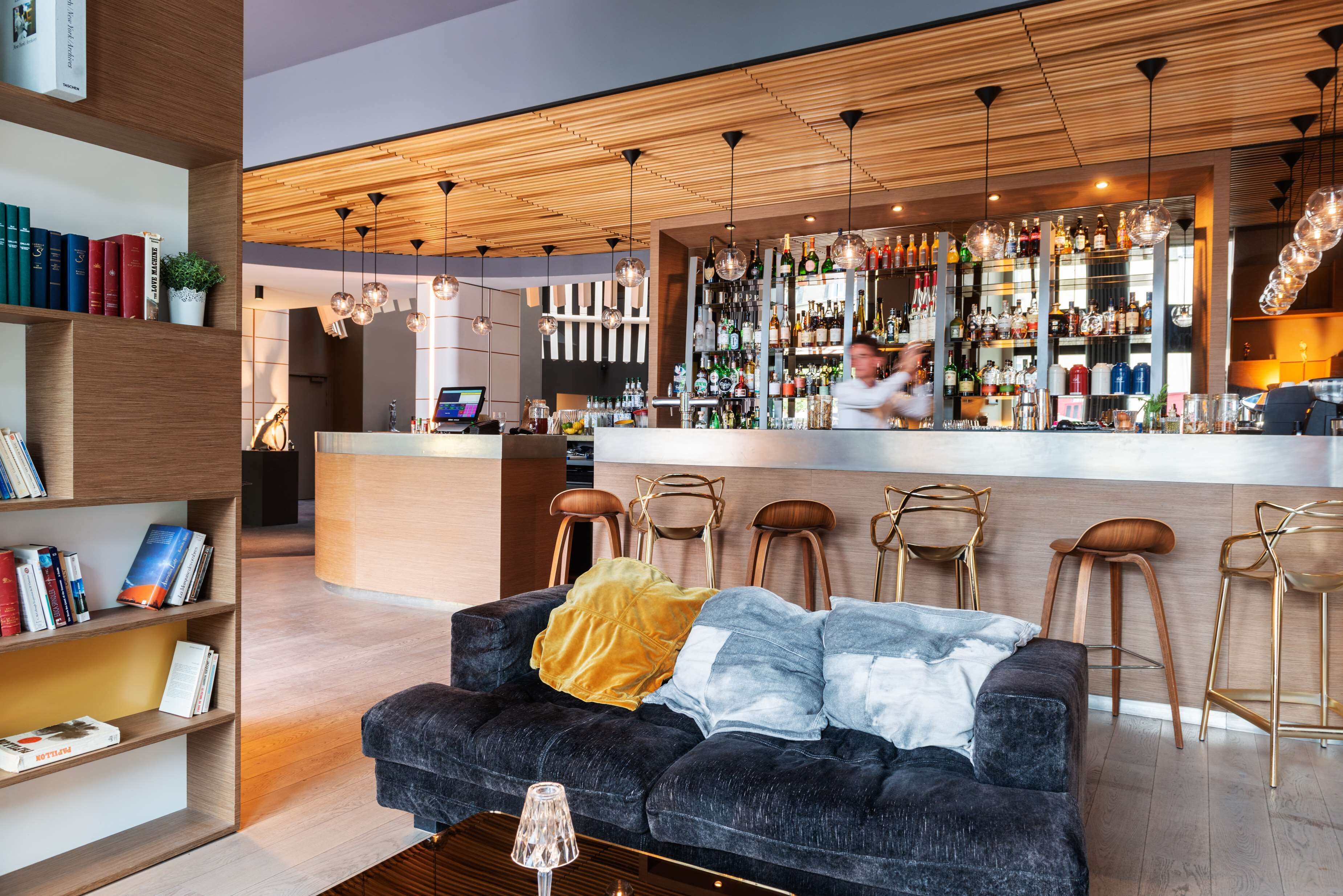 Un espace bar moderne, doté d’un comptoir, d’étagères remplies de bouteilles et de luminaires suspendus.