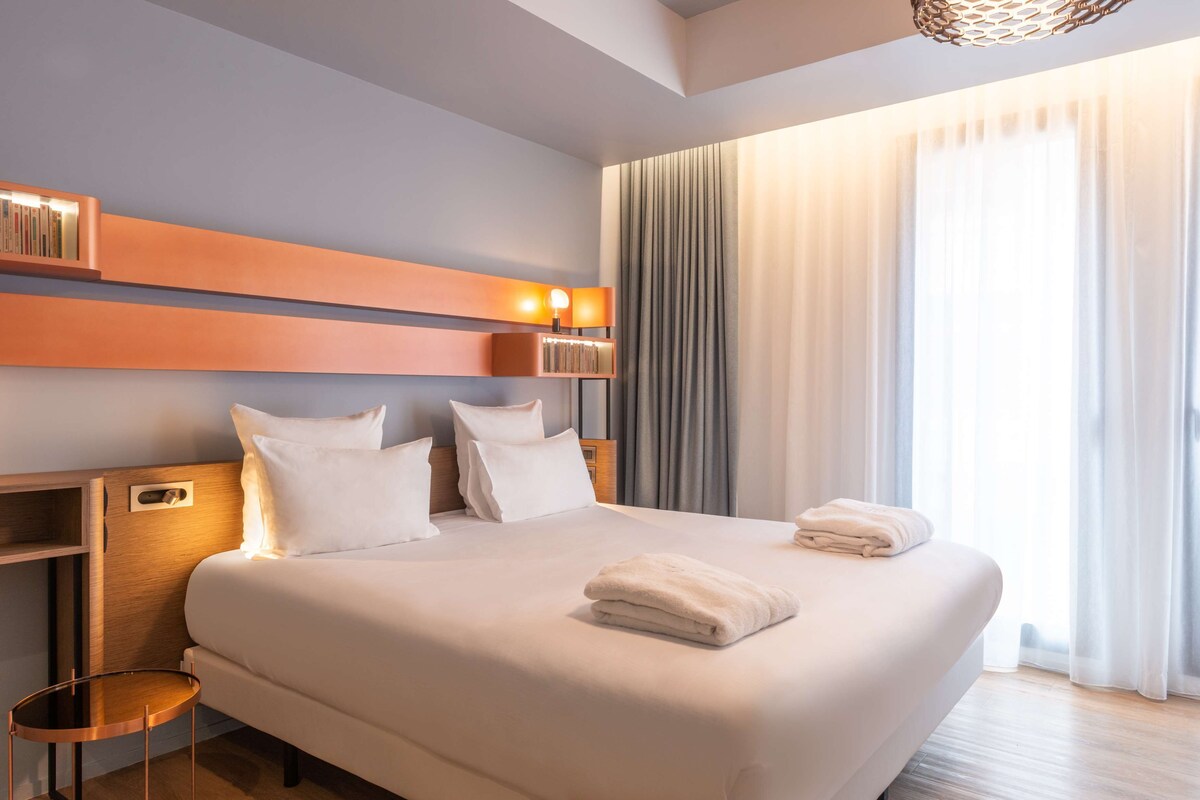 Ein modernes Hotelzimmer mit einem großen Bett, einem Nachttisch, einer eingebauten Bücherablage und einem großen Fenster mit Vorhängen.