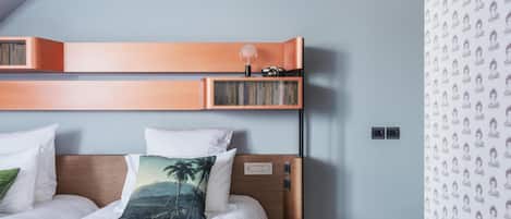 A hotel room with a bed, pillows, a side table, a lamp, and a bookshelf.