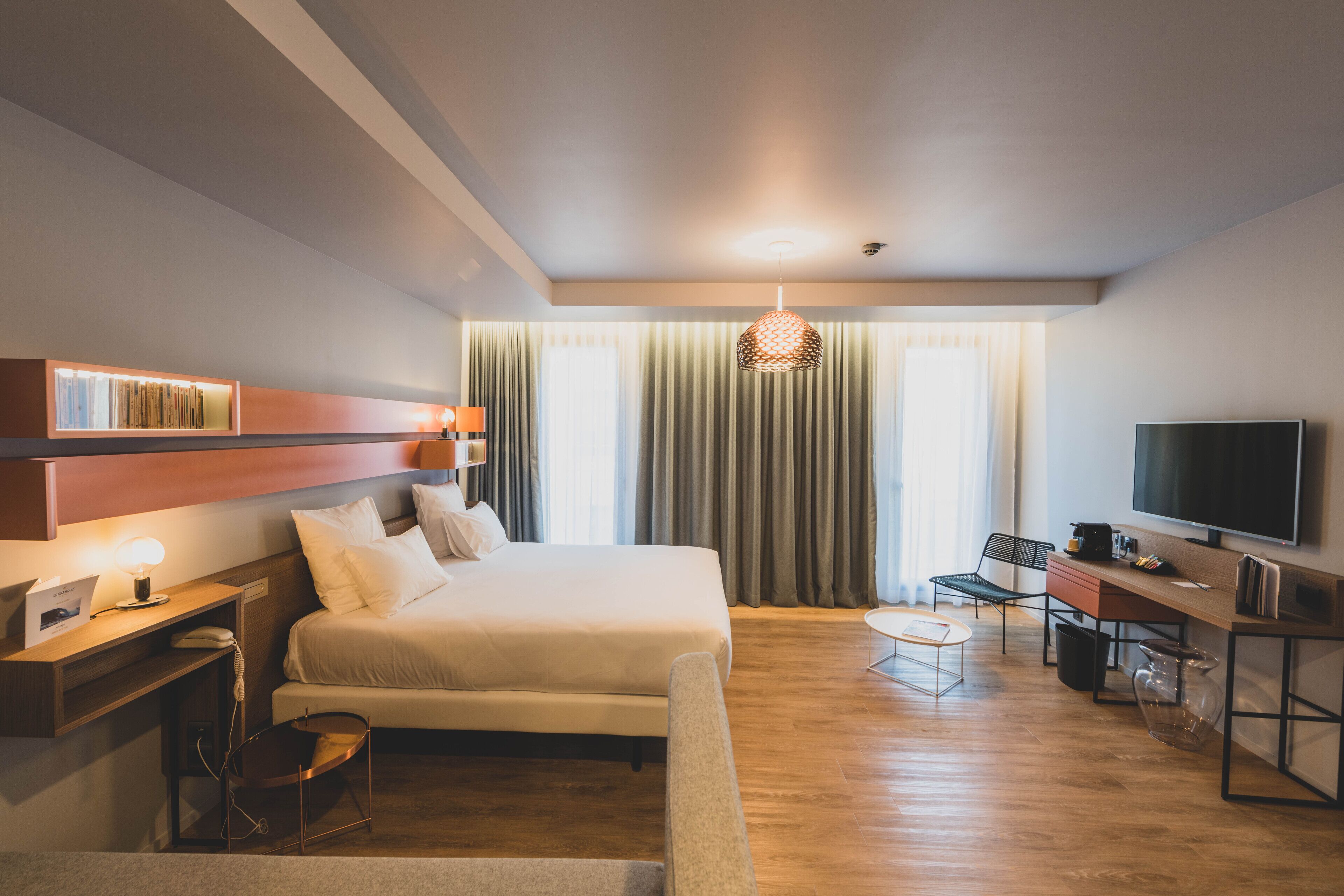 A modern hotel room with a bed, desk, chair, TV, and a large window with curtains.