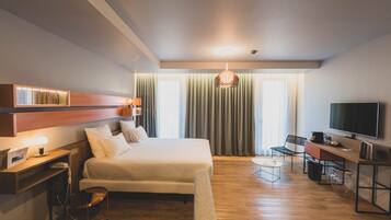 Een moderne hotelkamer met een bed, bureau, stoel, televisie en een groot raam met gordijnen.