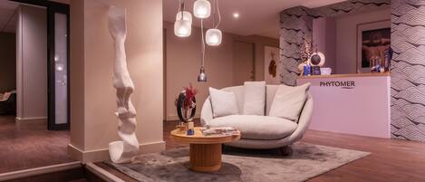Un vestíbulo moderno con una recepción que dice 'PHYTOMER', un sofá blanco, una mesa de centro de madera con revistas y una escultura blanca única.