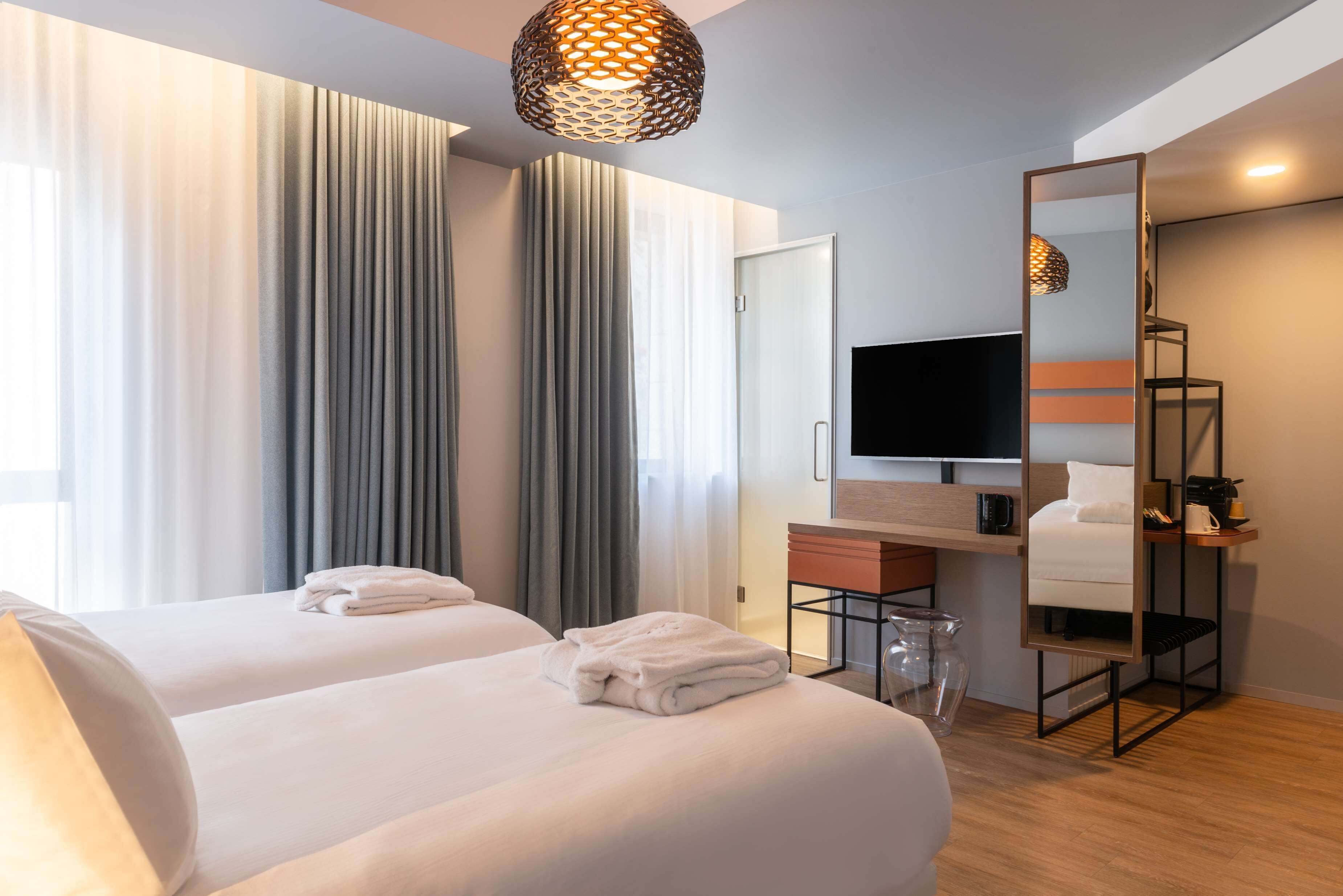 A hotel room with two beds, a TV, a mirror, and a large window with curtains.
