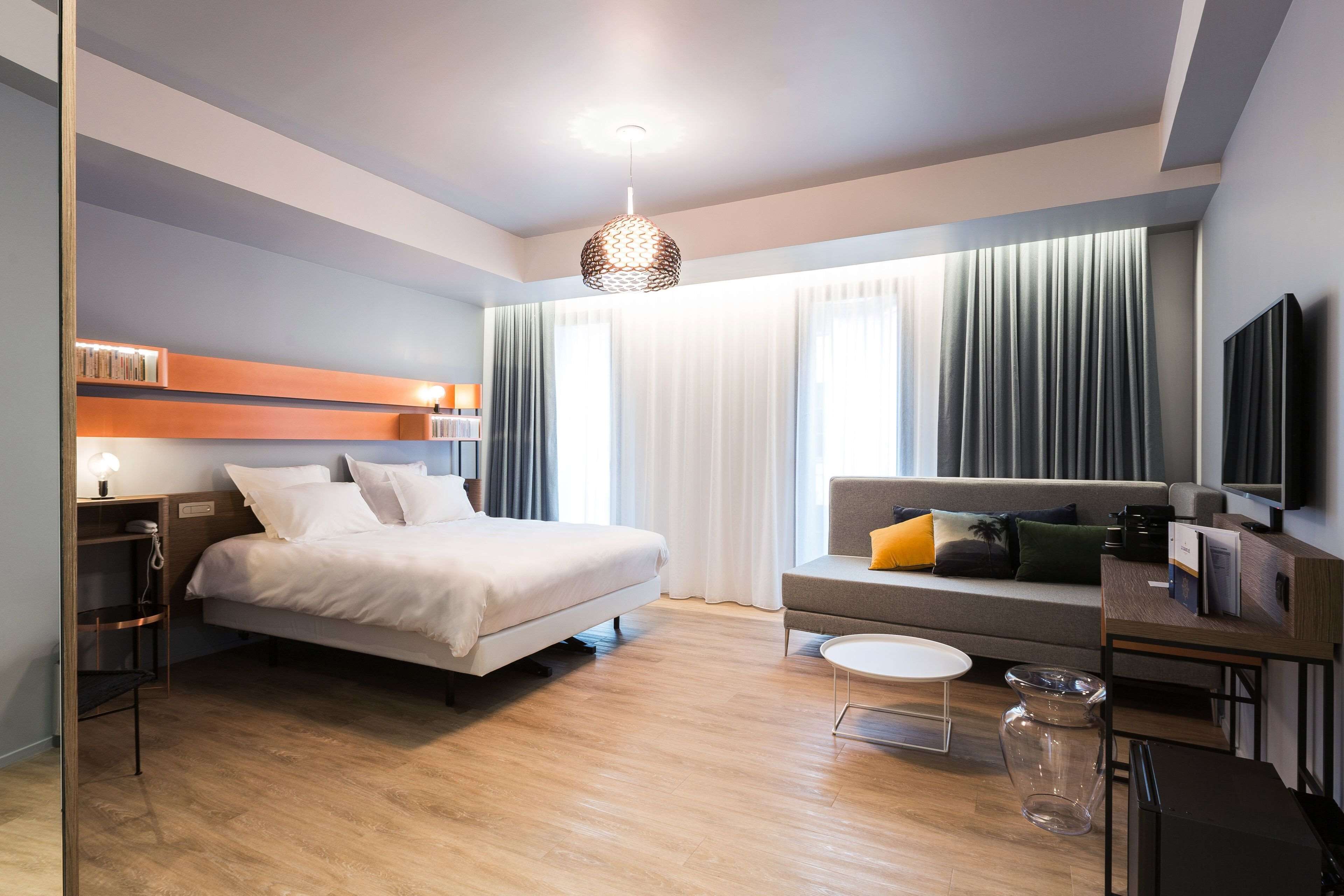 A modern hotel room with a bed, a sofa, a TV, and a desk with a lamp.