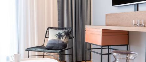 A modern hotel room with a wooden desk, a chair with a palm tree cushion, a small round table with a white mug, and a large TV mounted on the wall.