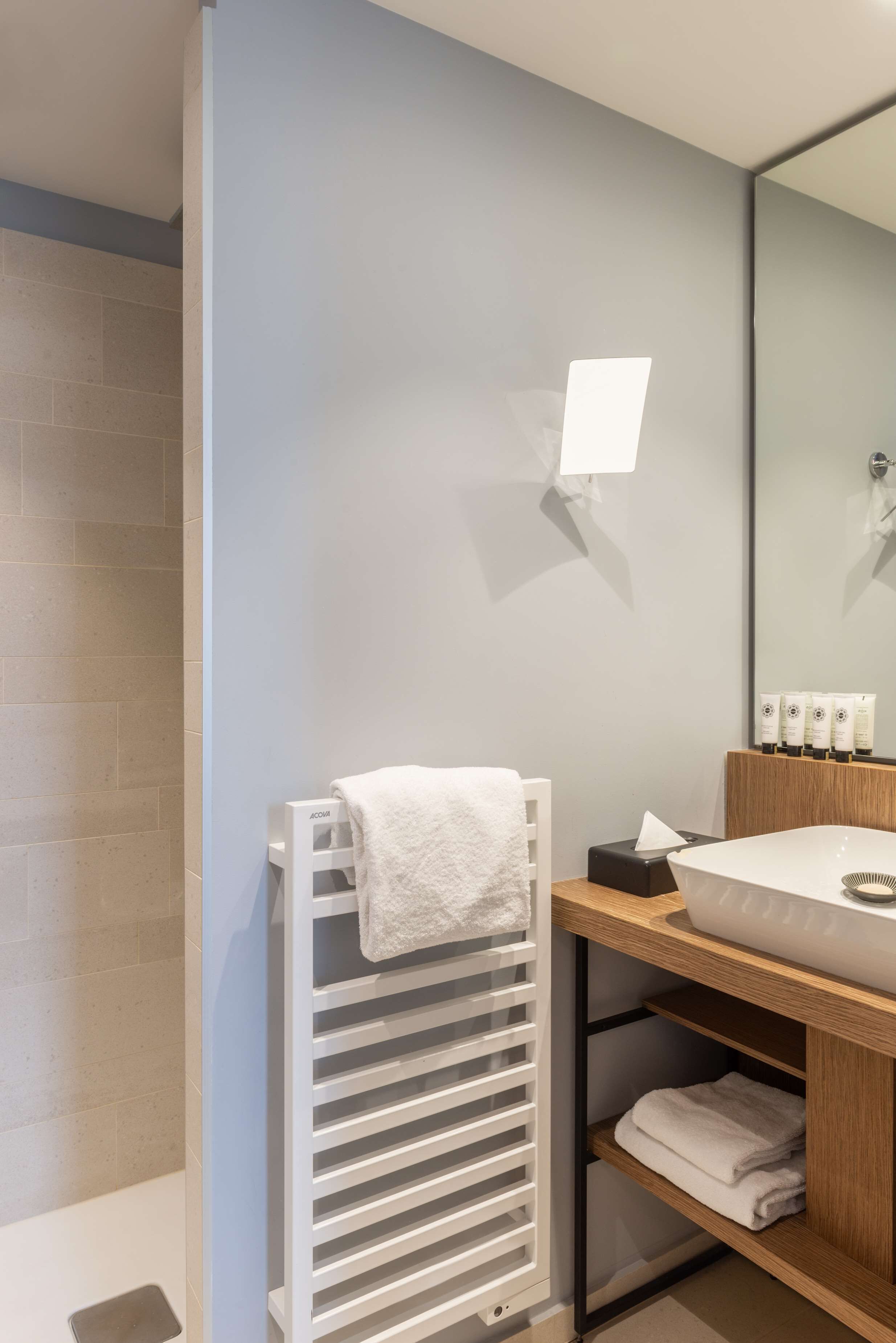 Un baño moderno con una toalla blanca en un perchero, un lavamanos con un espejo encima y artículos de aseo en un estante.