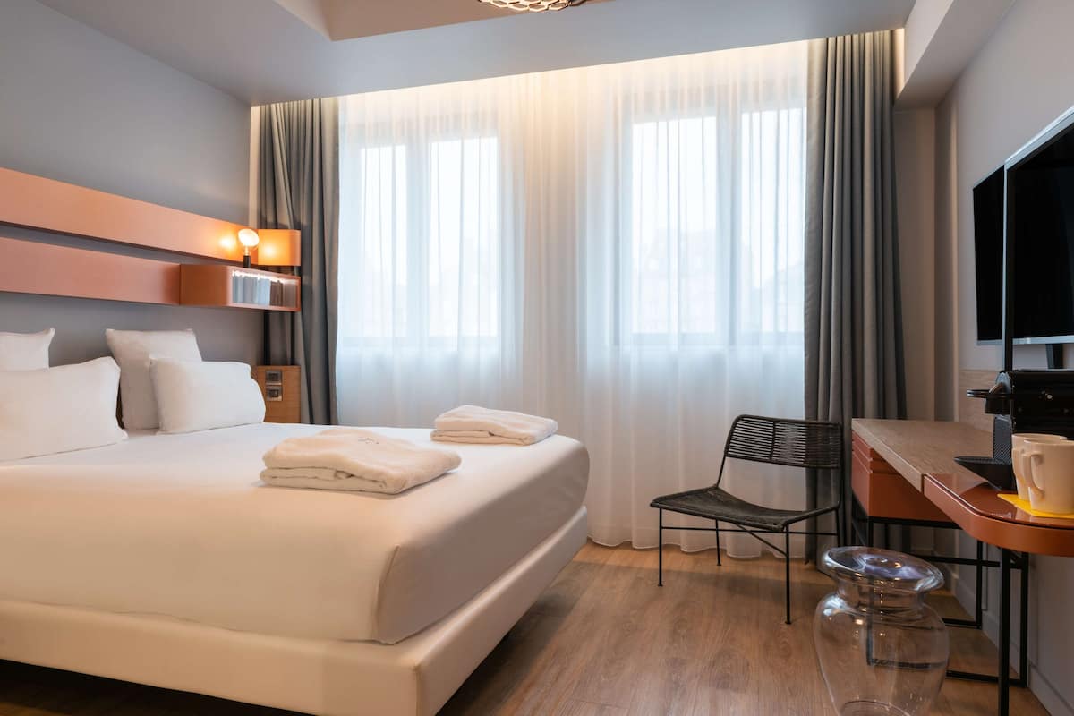 Ein Hotelzimmer mit Bett, Schreibtisch, Stuhl, Fernseher und einem Fenster mit Vorhängen.