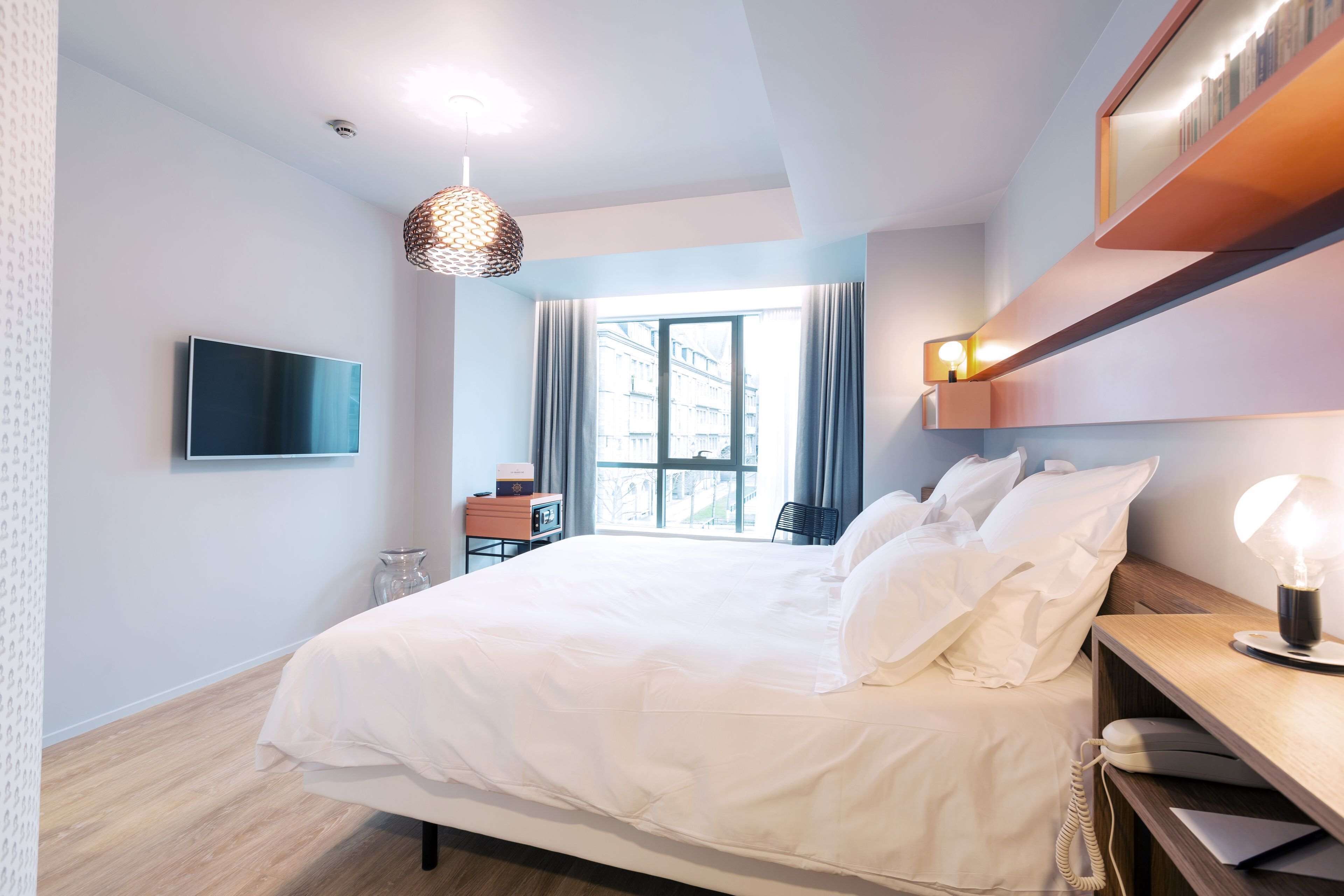 Habitación de hotel moderna con una cama grande, un televisor de pantalla plana, una mesita de noche con lámpara y vistas a la ciudad a través de un ventanal amplio.